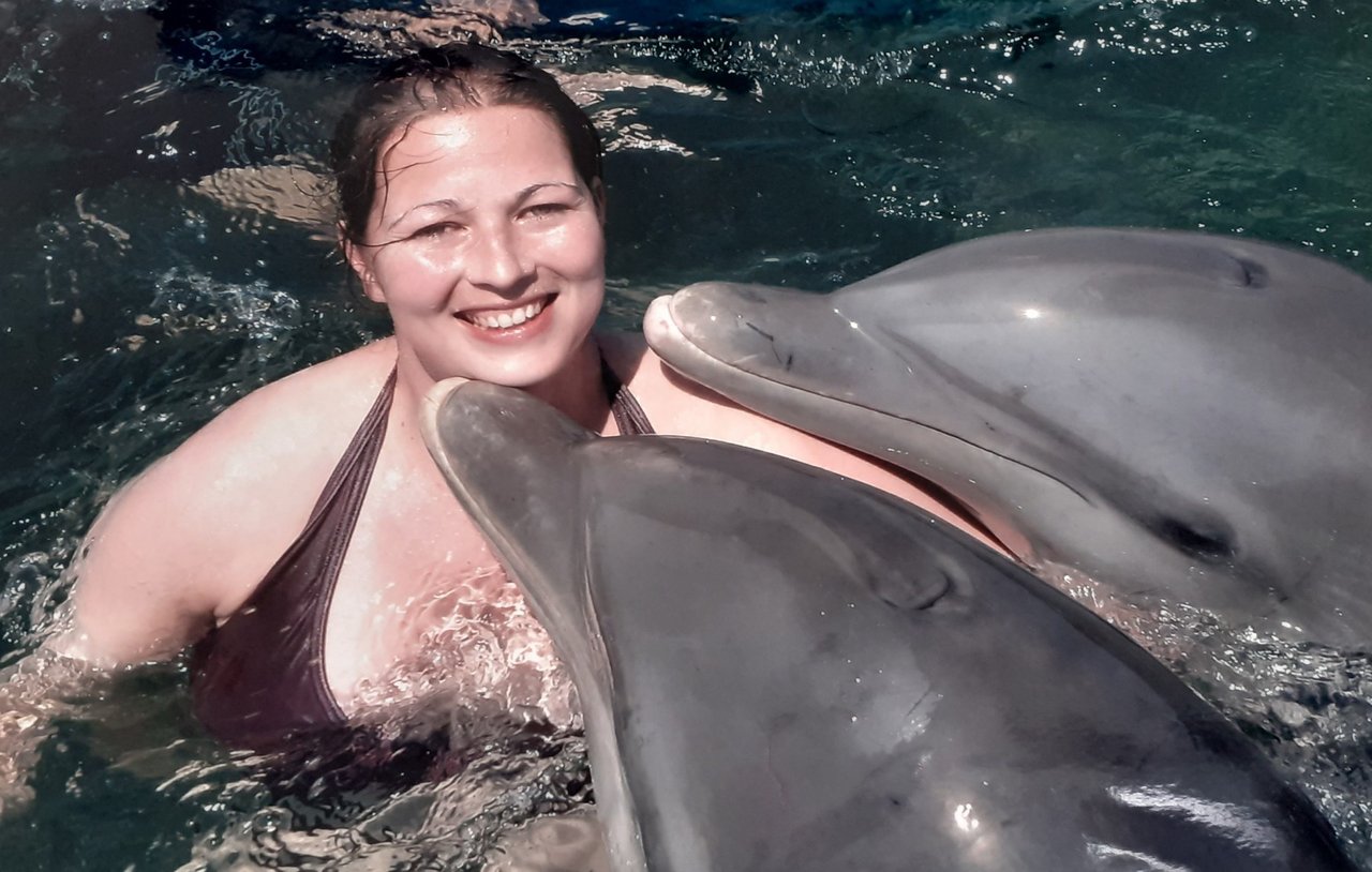 Mulher debruçada no braço de um barco, que está em alto mar, observando golfinhos que nadam ao lado da embarcação.